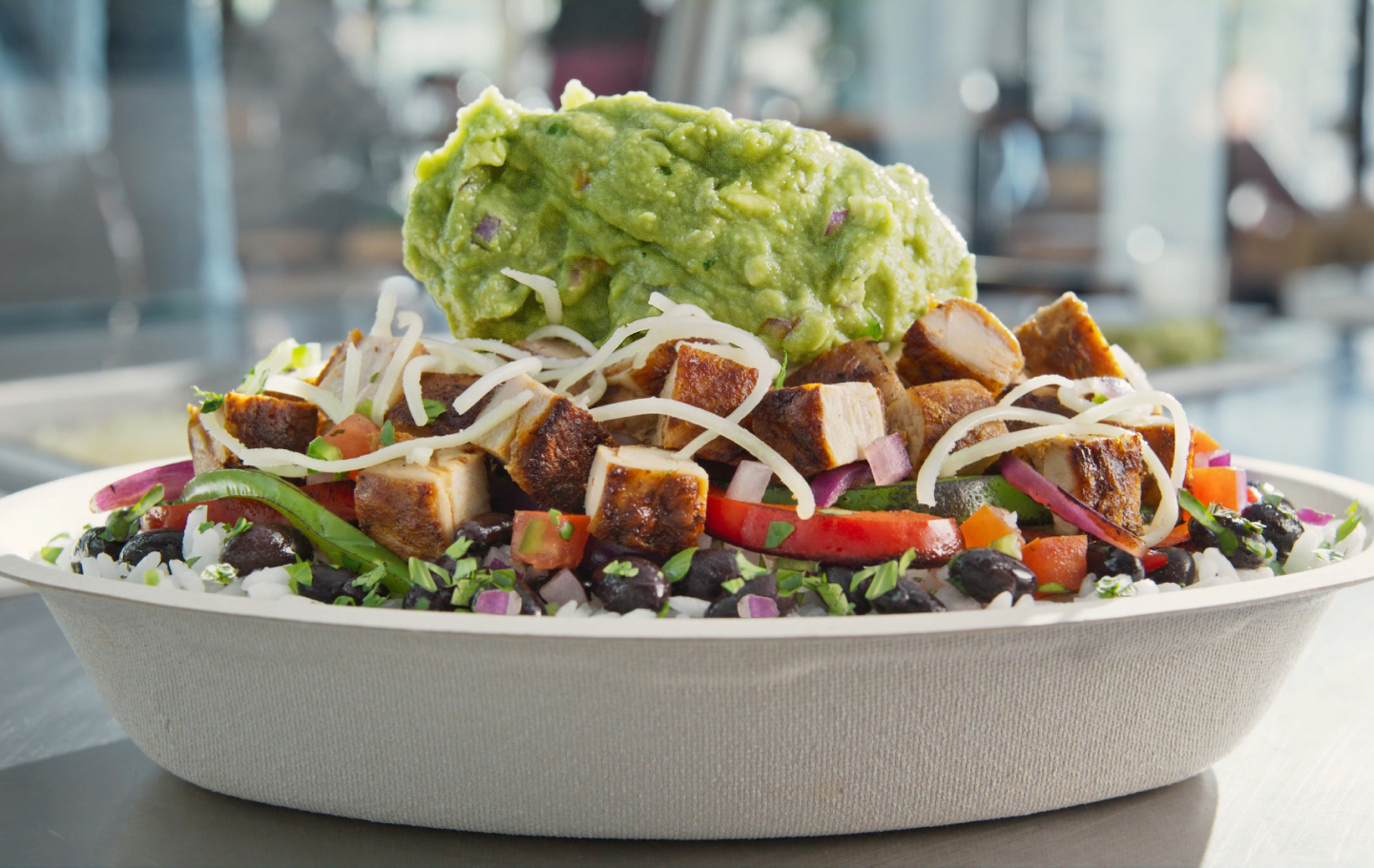 A Chipotle Burrito Bowl with black beans, fajita veggies, adobo chicken, fresh tomato salsa, monterey jack shredded cheese, being topped with hand-mashed guac.